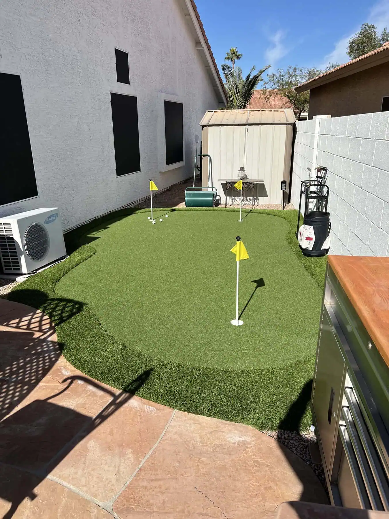 A small backyard golfer’s dream: a custom putting green with three golf holes and flags, surrounded by artificial grass golf green, next to a white house, storage shed, and various outdoor equipment.