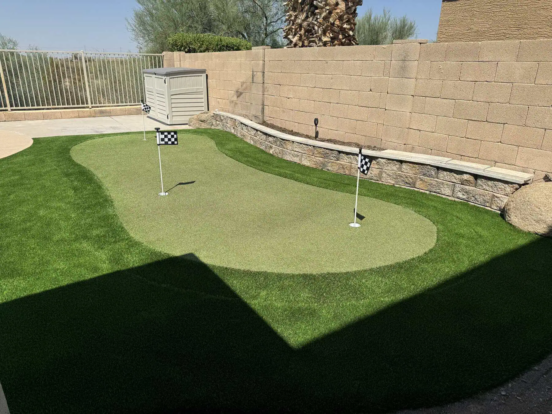 A small backyard golfer's paradise featuring a custom putting green with two checkered flags, surrounded by artificial turf, stone edging, and a tan brick wall under sunny skies.