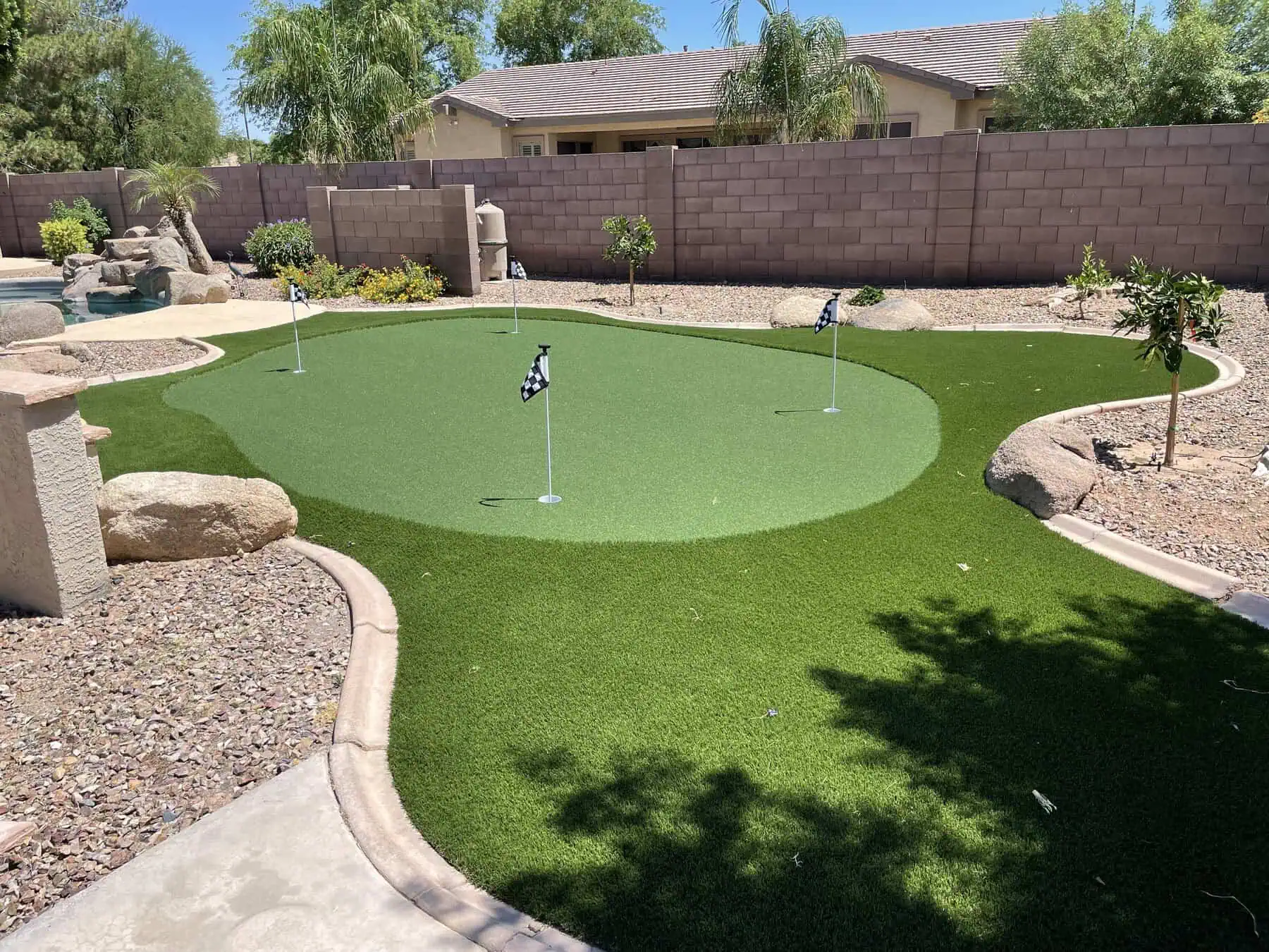 A backyard putting green for perfecting your short game, featuring three golf flags, surrounded by artificial grass installation, desert landscaping with palm trees, rocks, and a block wall in the background.