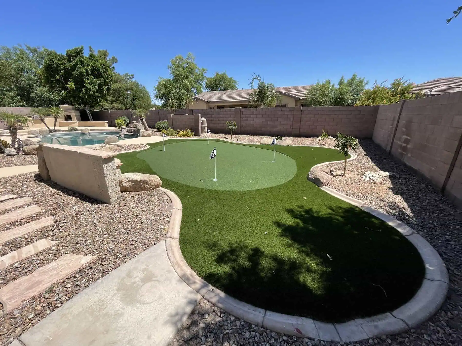 A backyard with a small artificial grass putting green for perfecting your short game, three flags, surrounding rocks, and desert landscaping. A hot tub and pool are on the left, with trees, a stone path nearby, and a block wall enclosing the yard.