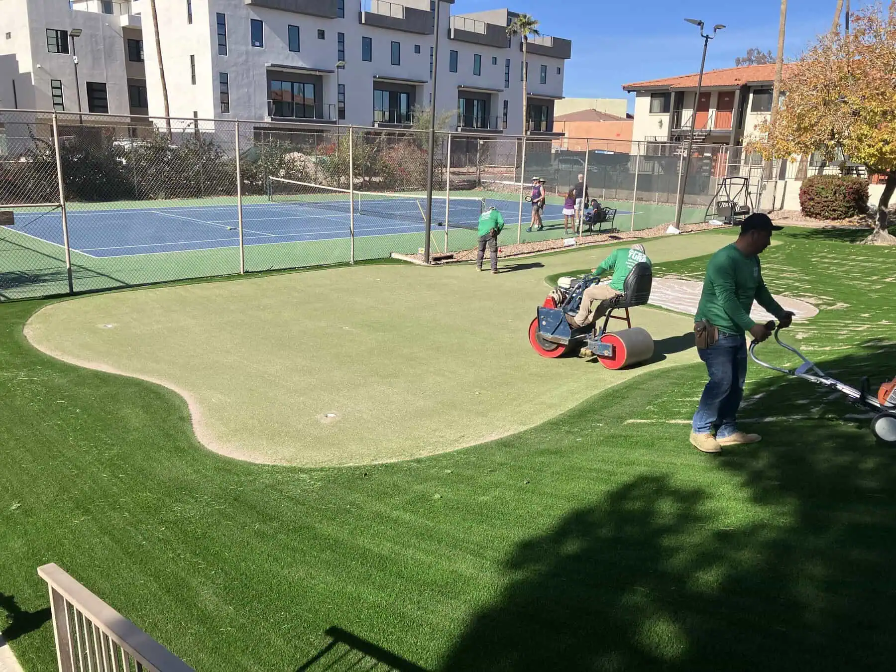 Workers install artificial turf around a custom putting green near a tennis court in a residential area, using lawn equipment and rollers on a sunny day.