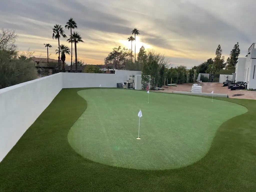 A custom-designed putting green with several golf holes and flags, surrounded by artificial turf and bordered by a white wall, sits beneath Phoenix palm trees with a sunset sky in the background.