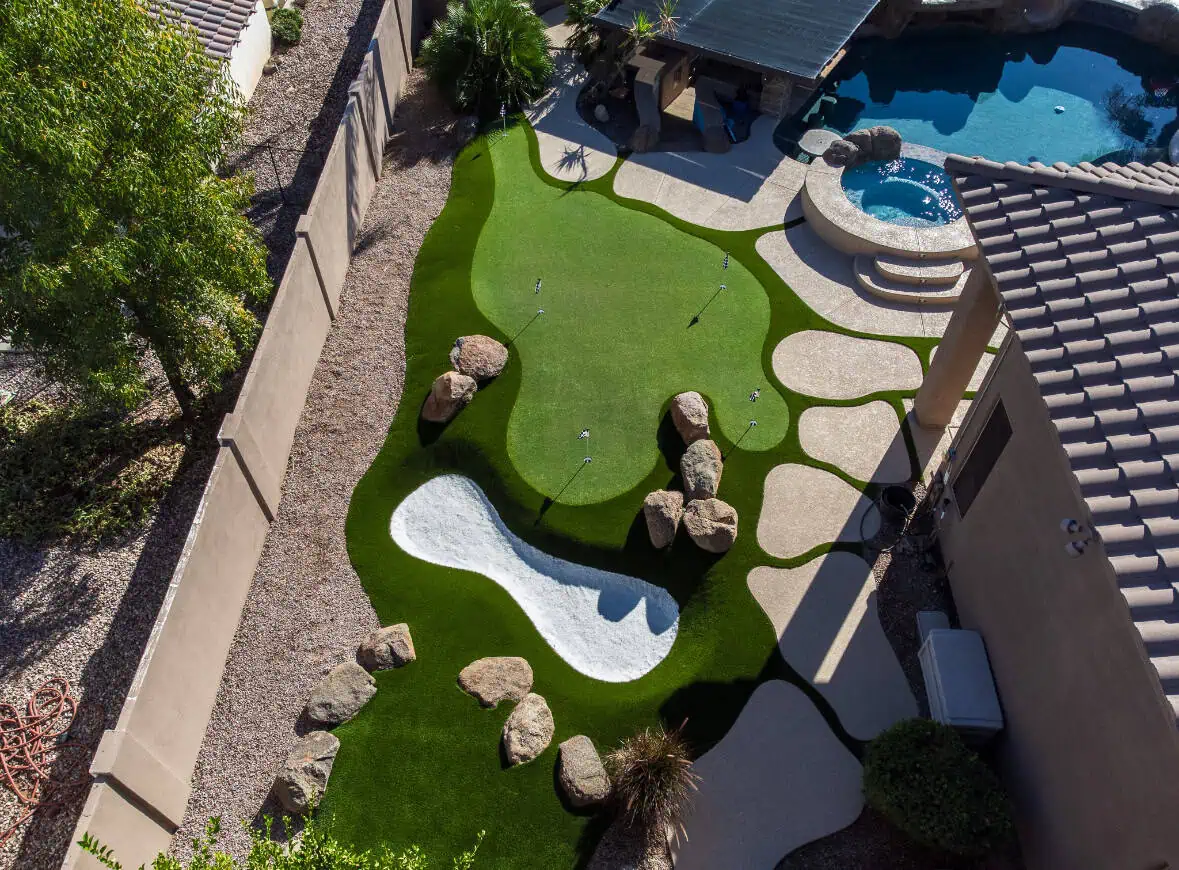 Aerial view of a Phoenix AZ backyard featuring a custom backyard putting green with artificial grass greens, sand trap with rocks, paved pathways, landscaped areas, and a swimming pool with hot tub beside a covered patio.
