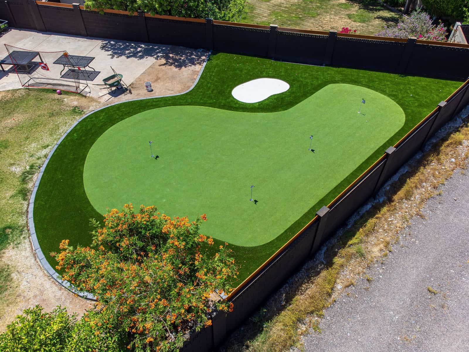 A backyard outdoor golf green features custom putting greens with artificial grass, a small white sand bunker, golf flags, and is surrounded by a fence and patio furniture nearby.