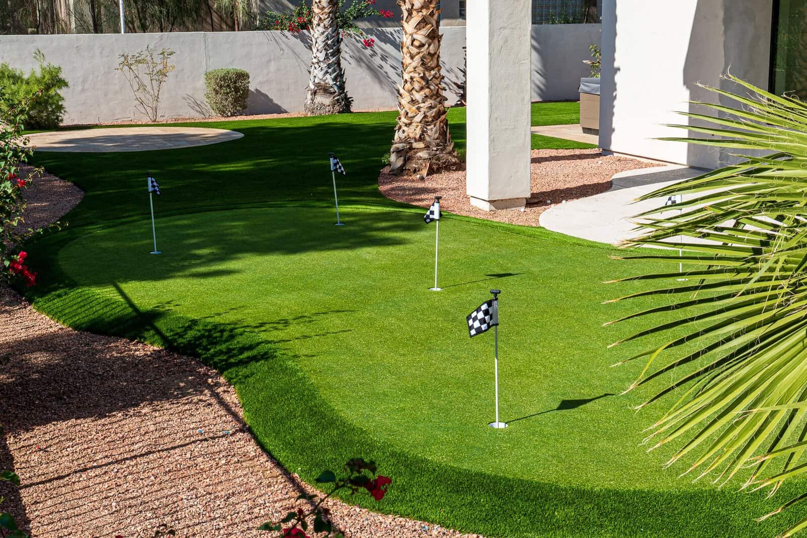 A backyard putting green with several small checkered flags marking holes, surrounded by lush artificial grass from expert turf installations, palm trees, and desert landscaping.
