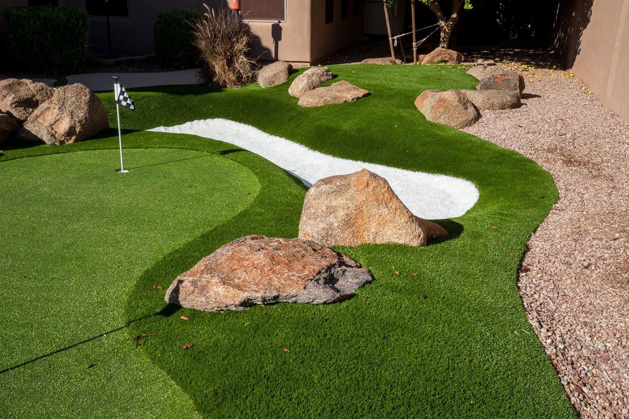 A small artificial putting green made from quality artificial grass with a white sand bunker, scattered rocks, and a checkered flag, surrounded by gravel and shrubs next to a building.