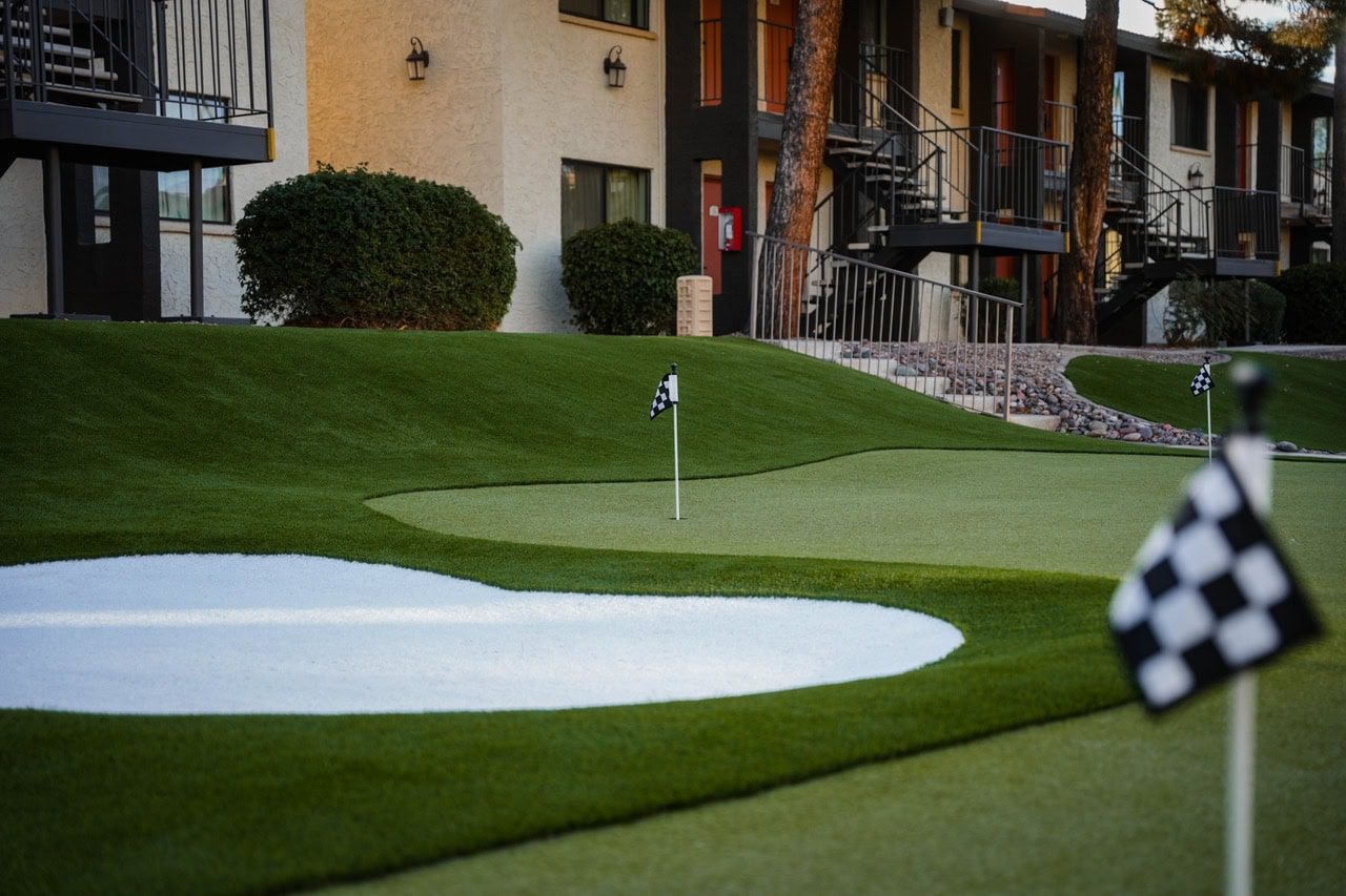 A custom putting green with artificial grass golf green, small checkered flags, and a sand trap is featured in the courtyard of an apartment complex with beige walls, balconies, and staircases. Shrubs line the building.