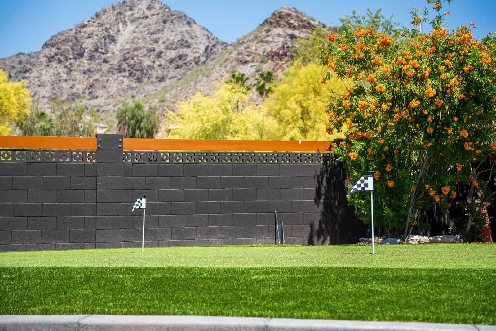 A small custom backyard putting green with golf flags is set in a Phoenix AZ yard, bordered by a black brick wall. Behind the artificial grass green are flowering trees and mountains under a clear blue sky.