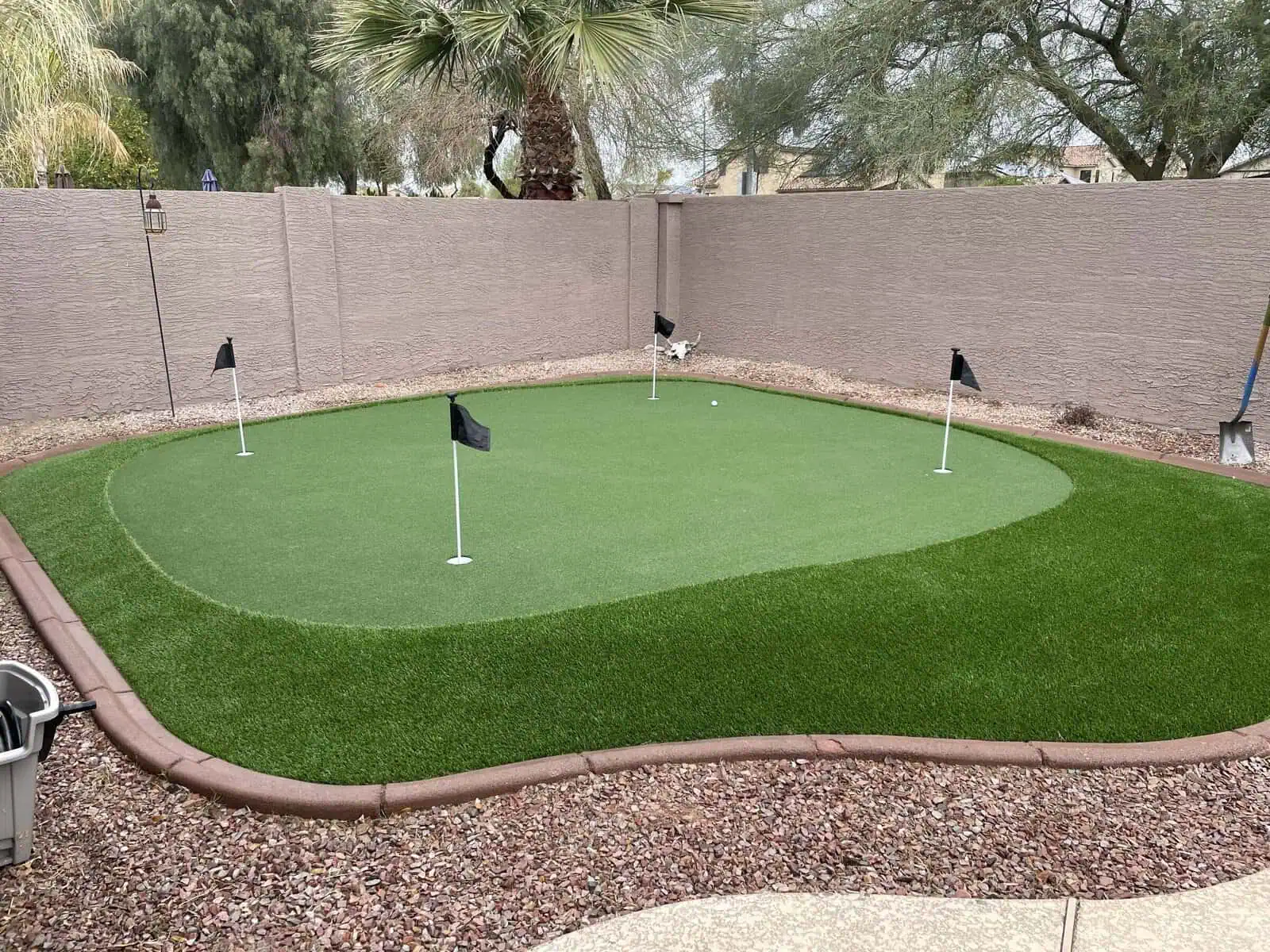 A backyard putting green with four holes, each marked by a small black flag, features expert artificial grass installation and is surrounded by landscaping rocks, palm trees, and beige privacy walls—perfect for improving your short game.