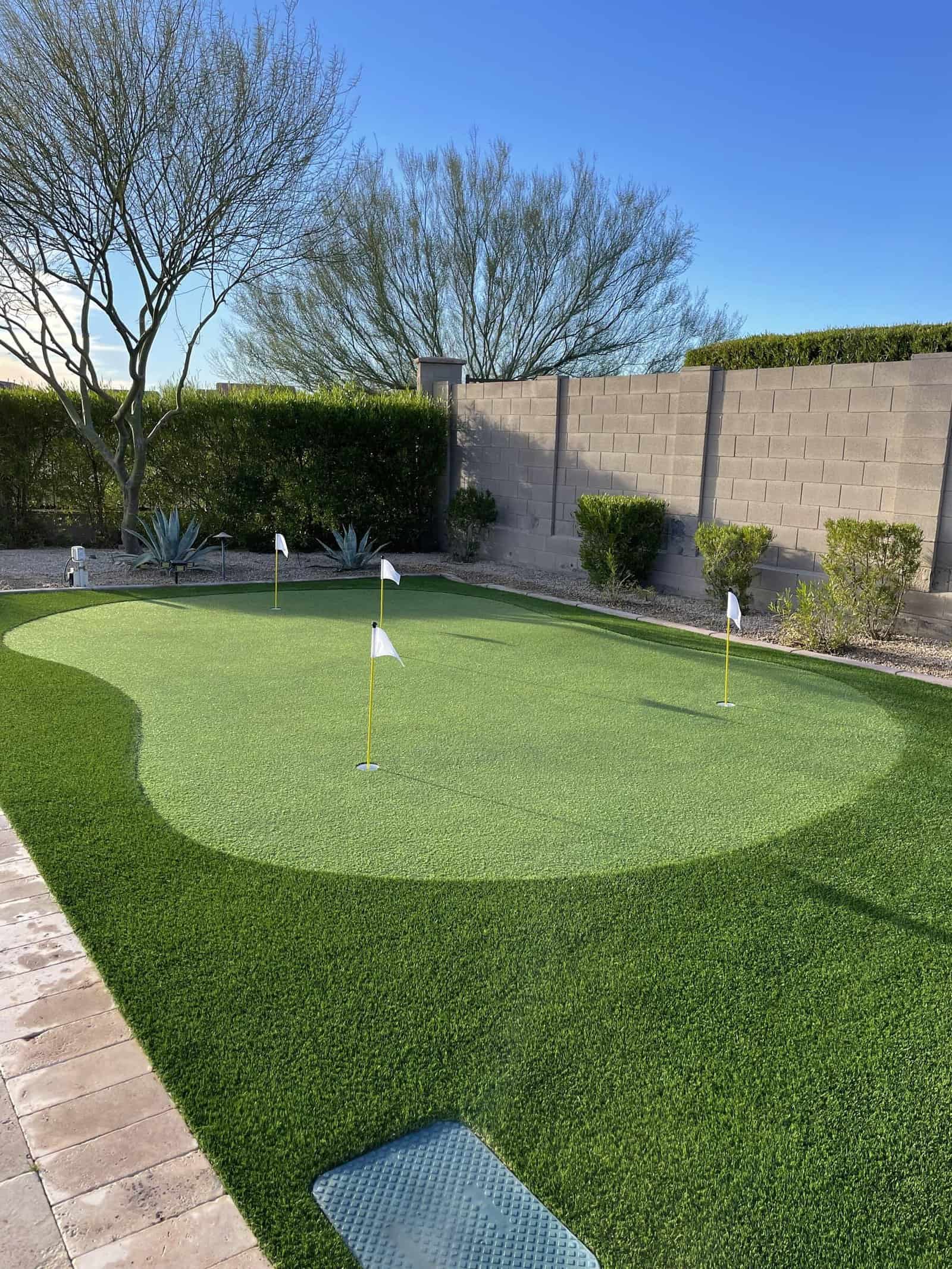 A backyard golf green with custom putting greens, featuring artificial turf, four small golf flags, desert landscaping, and a stone wall fence under a clear blue sky.