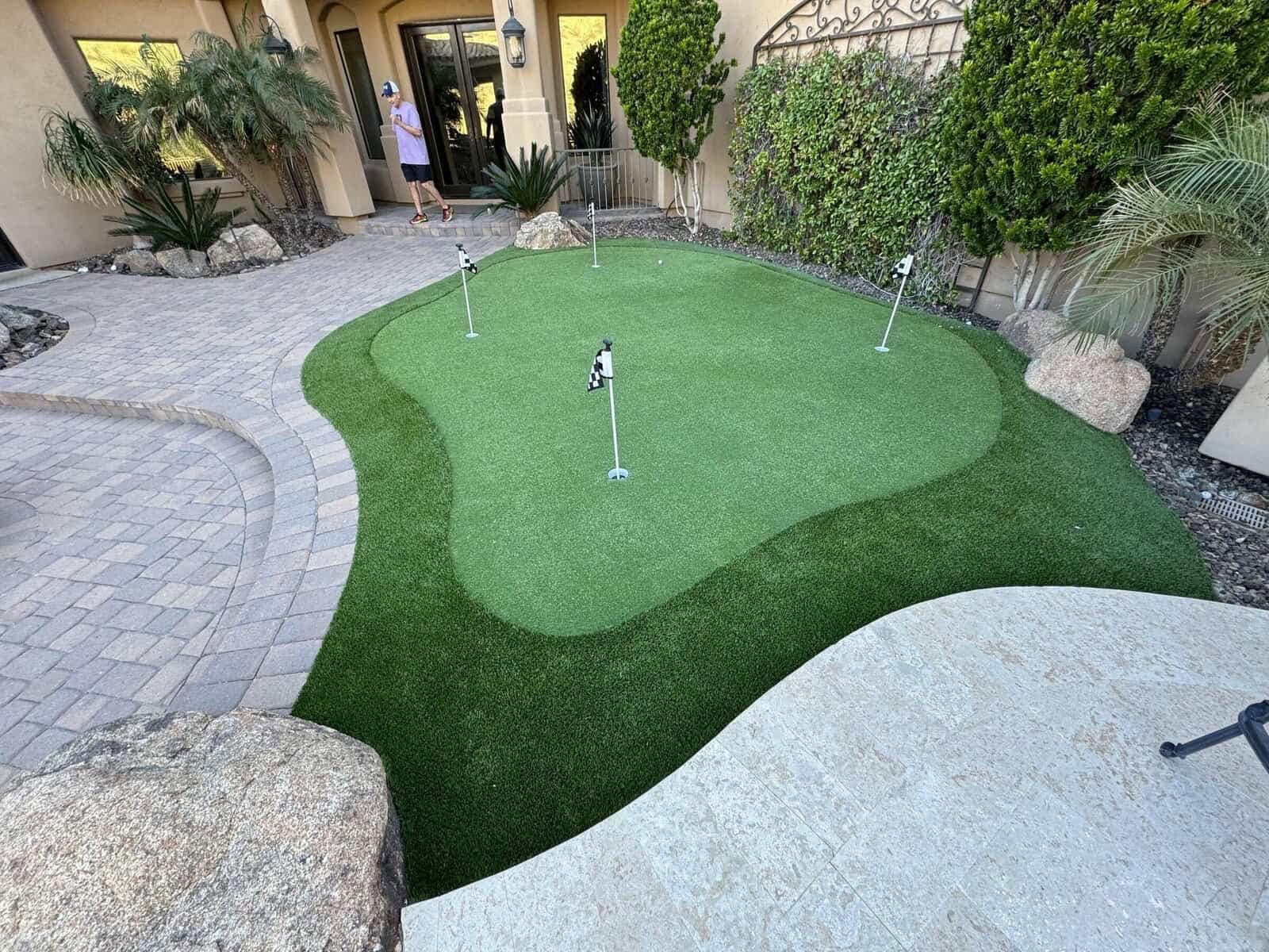 A small backyard putting green with artificial grass and four holes is surrounded by stone pavers, rocks, and lush greenery in a landscaped courtyard near a building in Phoenix AZ. A person stands near the entrance in the background.