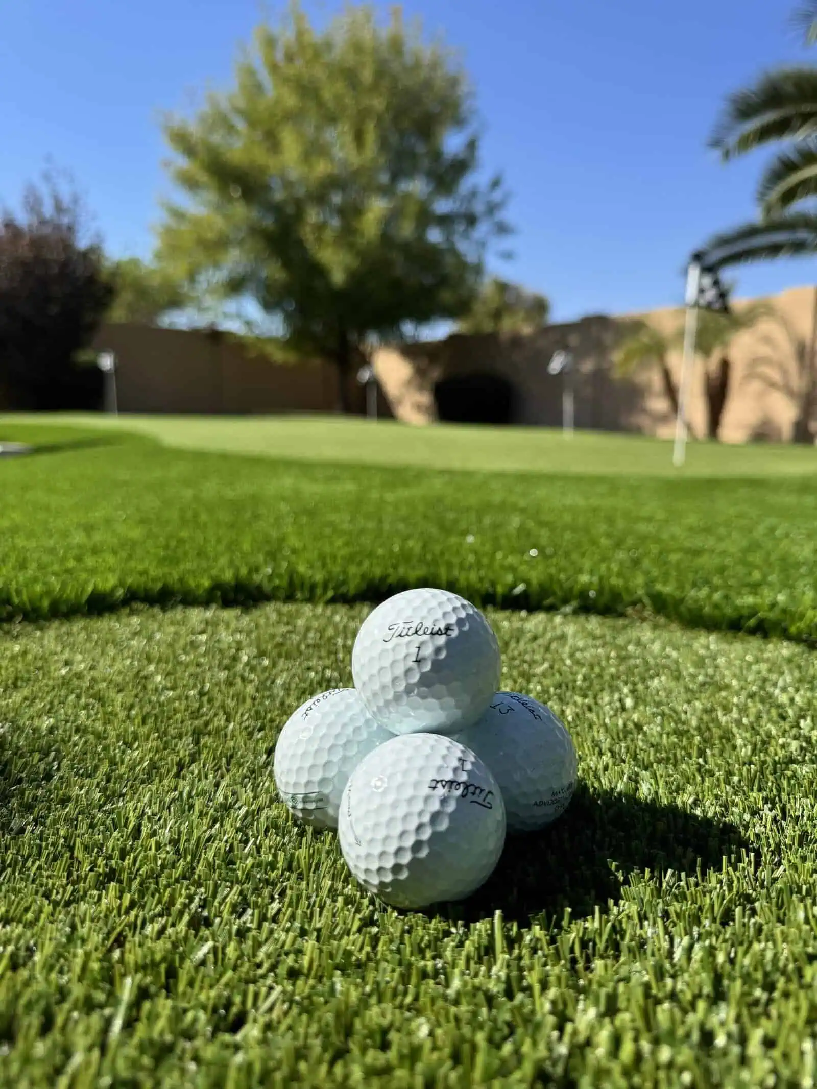 Four white golf balls stacked on lush artificial grass at a golf green, with trees, a sand-colored wall, and blue sky behind. A white flag marks the hole in the distance on this stunning custom putting green.