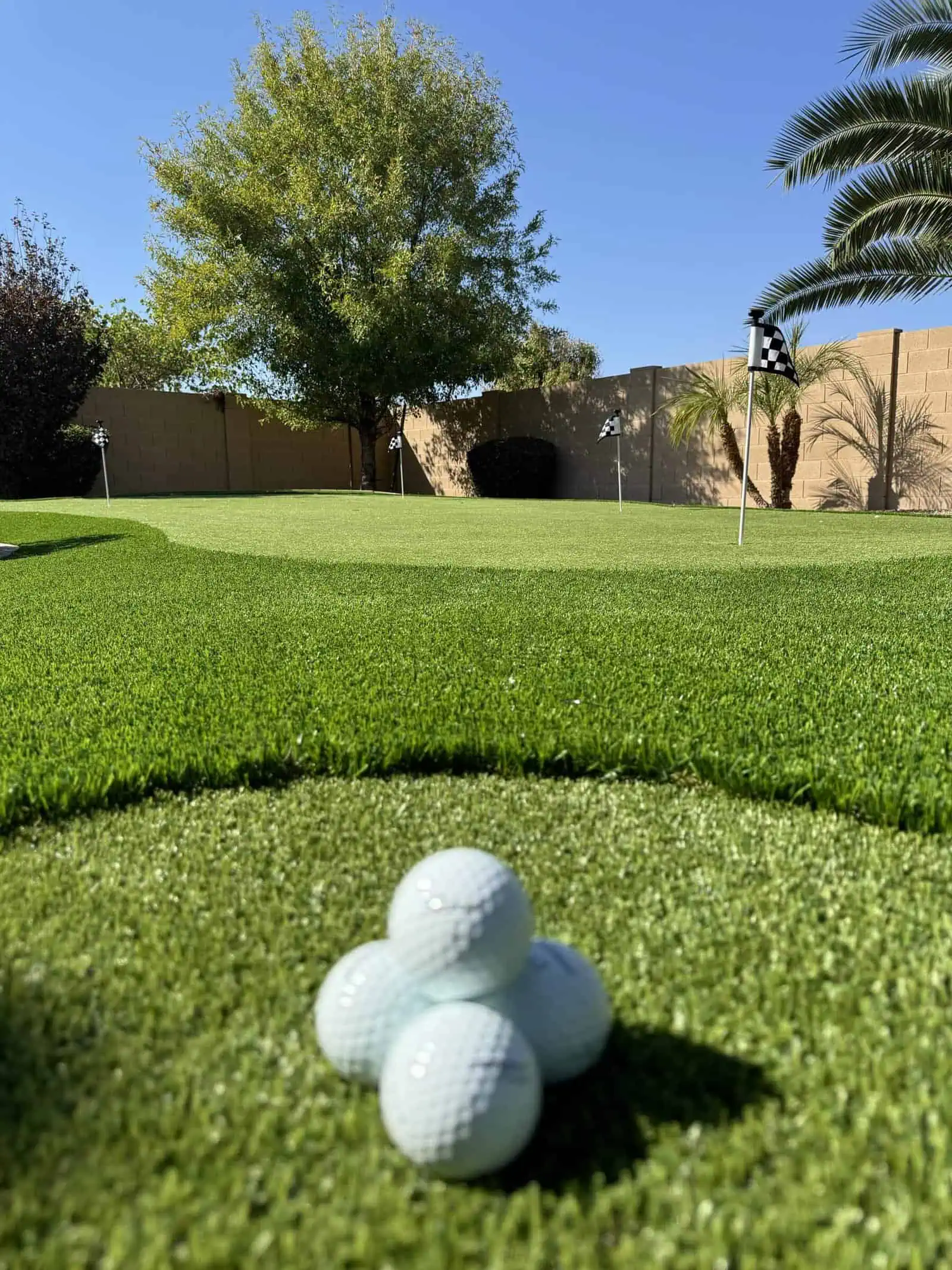 Four golf balls are stacked in a pyramid on artificial turf in the foreground, with an outdoor putting green, golf flags, palm trees, and a brick wall enhancing this sunny backyard’s custom putting greens setup.