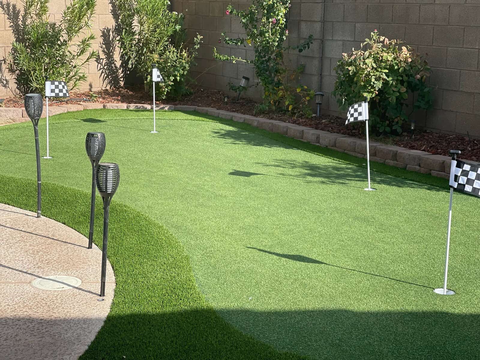A backyard putting green with artificial grass installation features four golf holes marked by checkered flags. Decorative torches line the edge, while plants and flowers flourish along a brick wall—a perfect spot to work on your short game.