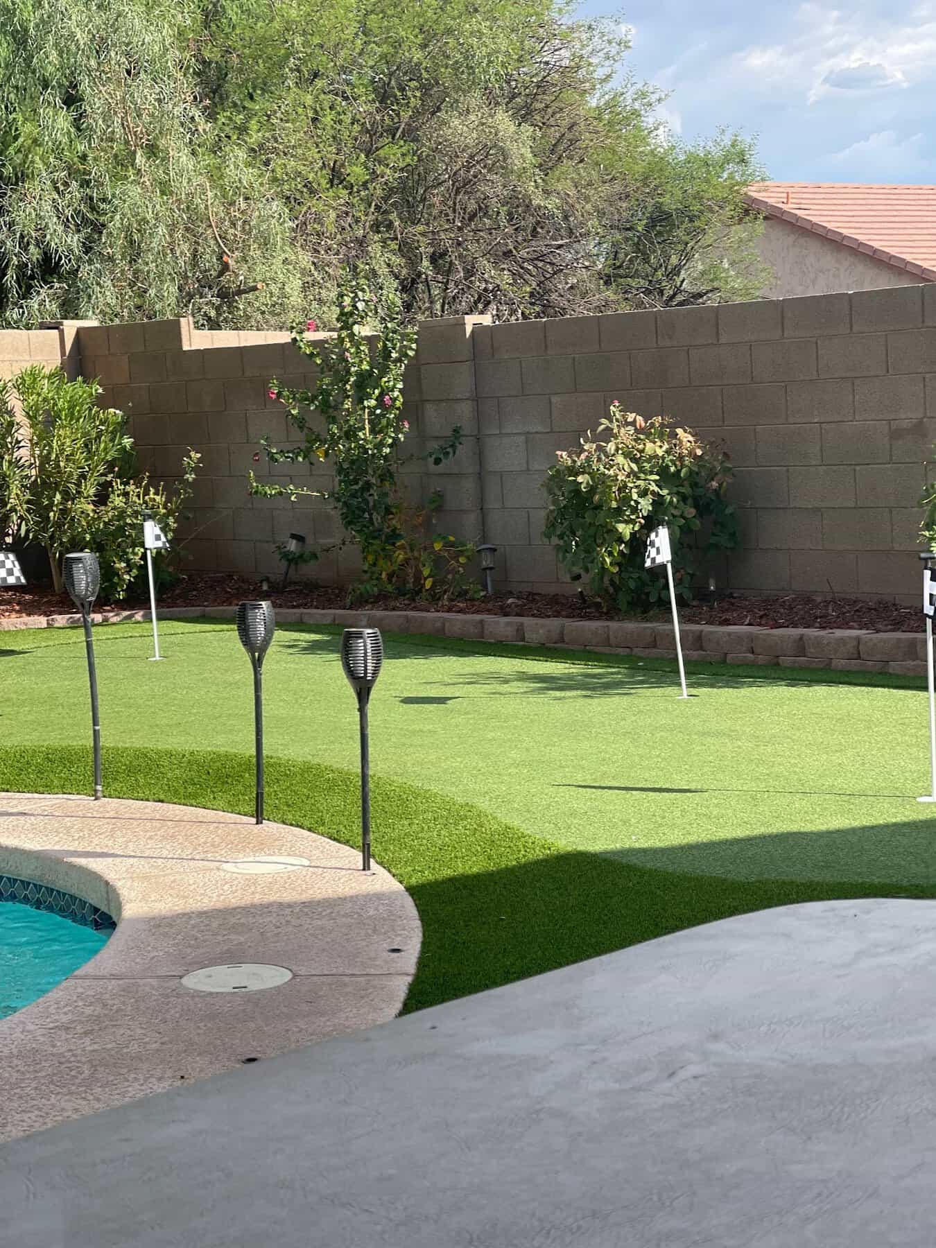 A Phoenix backyard features custom putting greens with four small golf flags, surrounded by plants and a block wall. Tiki torches line the edge of a curved pool in the foreground, creating a relaxing oasis.