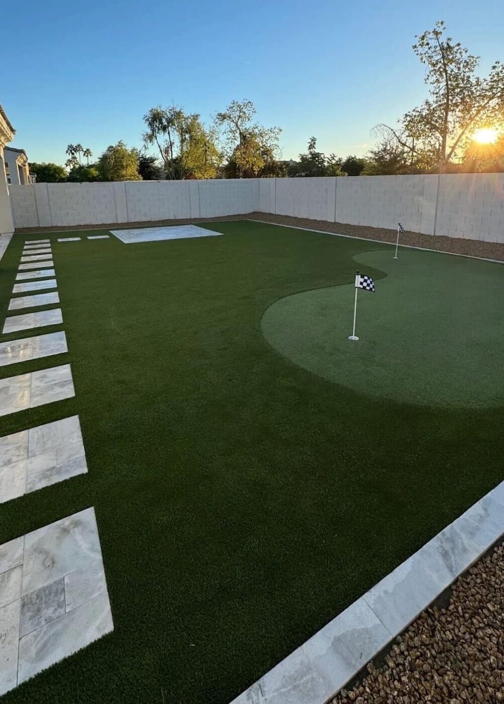 A Phoenix backyard features a large artificial grass lawn with a custom-designed putting green, marked by a checkered flag, bordered by stone pavers and a concrete patio, fenced in and surrounded by trees at sunset.