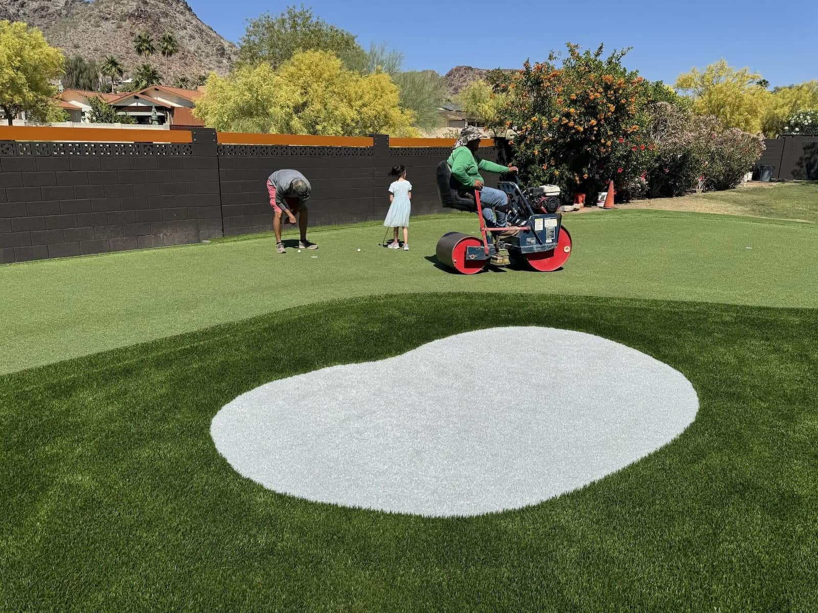 Three people enjoy a backyard putting green: one lines up a putt, a child stands nearby, and another rides a red lawn roller near the sand trap. Artificial grass greens recreate the feel of Phoenix AZ golf with scenic trees and mountains in the background.